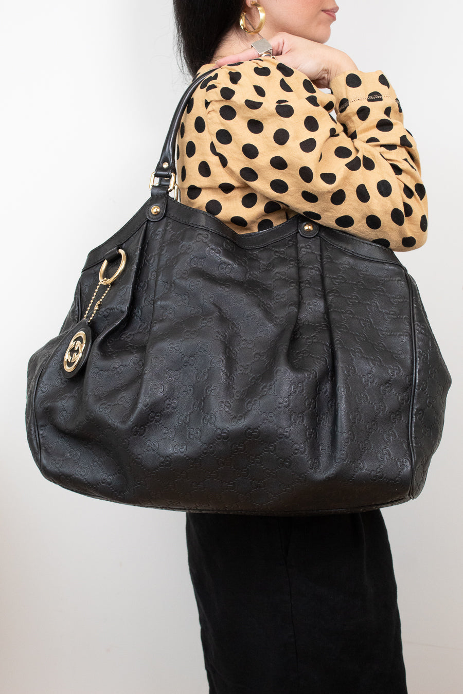 A person wearing a beige and black polka-dot top carries a large black leather shoulder bag with gold-tone hardware and a circular interlocking G logo charm. The person is also wearing black pants and gold hoop earrings, photographed against a plain light gray background.