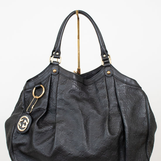 A black leather shoulder bag with an embossed monogram pattern, gold-tone hardware, and a hanging interlocking G logo charm, displayed on a white pedestal against a white background.