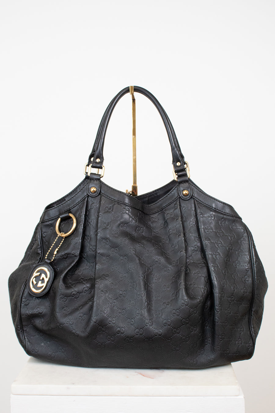 A black leather shoulder bag with an embossed monogram pattern, gold-tone hardware, and a hanging interlocking G logo charm, displayed on a white pedestal against a white background.