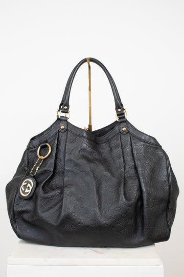 A black leather shoulder bag with an embossed monogram pattern, gold-tone hardware, and a hanging interlocking G logo charm, displayed on a white pedestal against a white background.