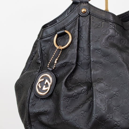 A black leather shoulder bag with an embossed monogram pattern, featuring gold-tone hardware, a hanging keychain with a Gucci logo patch, and a gold ring detail, shown against a plain white background.