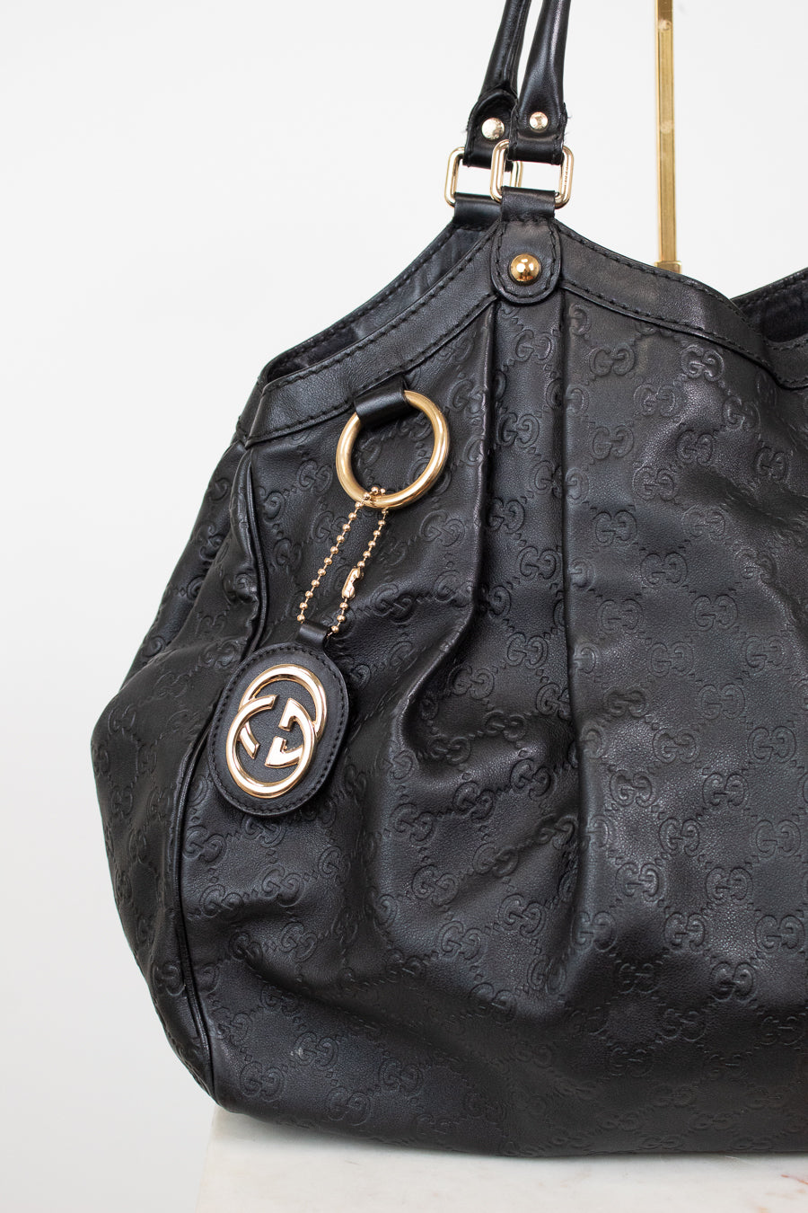 A black leather shoulder bag with an embossed monogram pattern, featuring gold-tone hardware, a hanging keychain with a Gucci logo patch, and a gold ring detail, shown against a plain white background.
