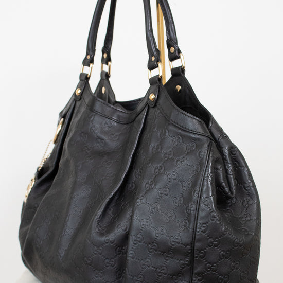 A black leather shoulder bag with an embossed monogram pattern, featuring gold-tone hardware and dual top handles, displayed on a white background.