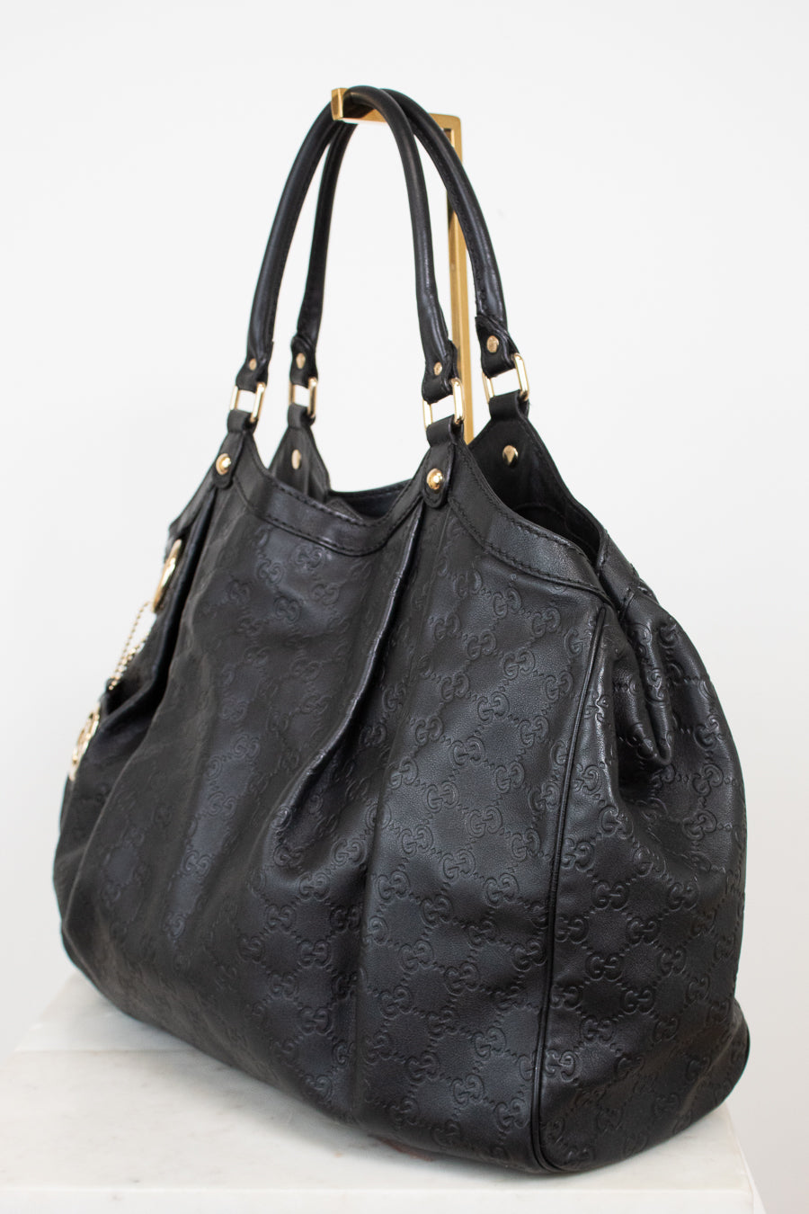 A black leather shoulder bag with an embossed monogram pattern, featuring gold-tone hardware and dual top handles, displayed on a white background.