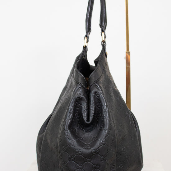 A black leather shoulder bag with embossed monogram pattern, displayed on a white surface with a gold metal stand against a white background.