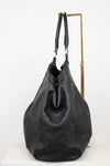 A black leather shoulder bag with embossed monogram pattern, displayed on a white surface with a gold metal stand against a white background.