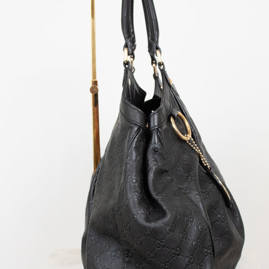 A black leather  handbag with an embossed monogram pattern, displayed on a gold metal stand against a white background.