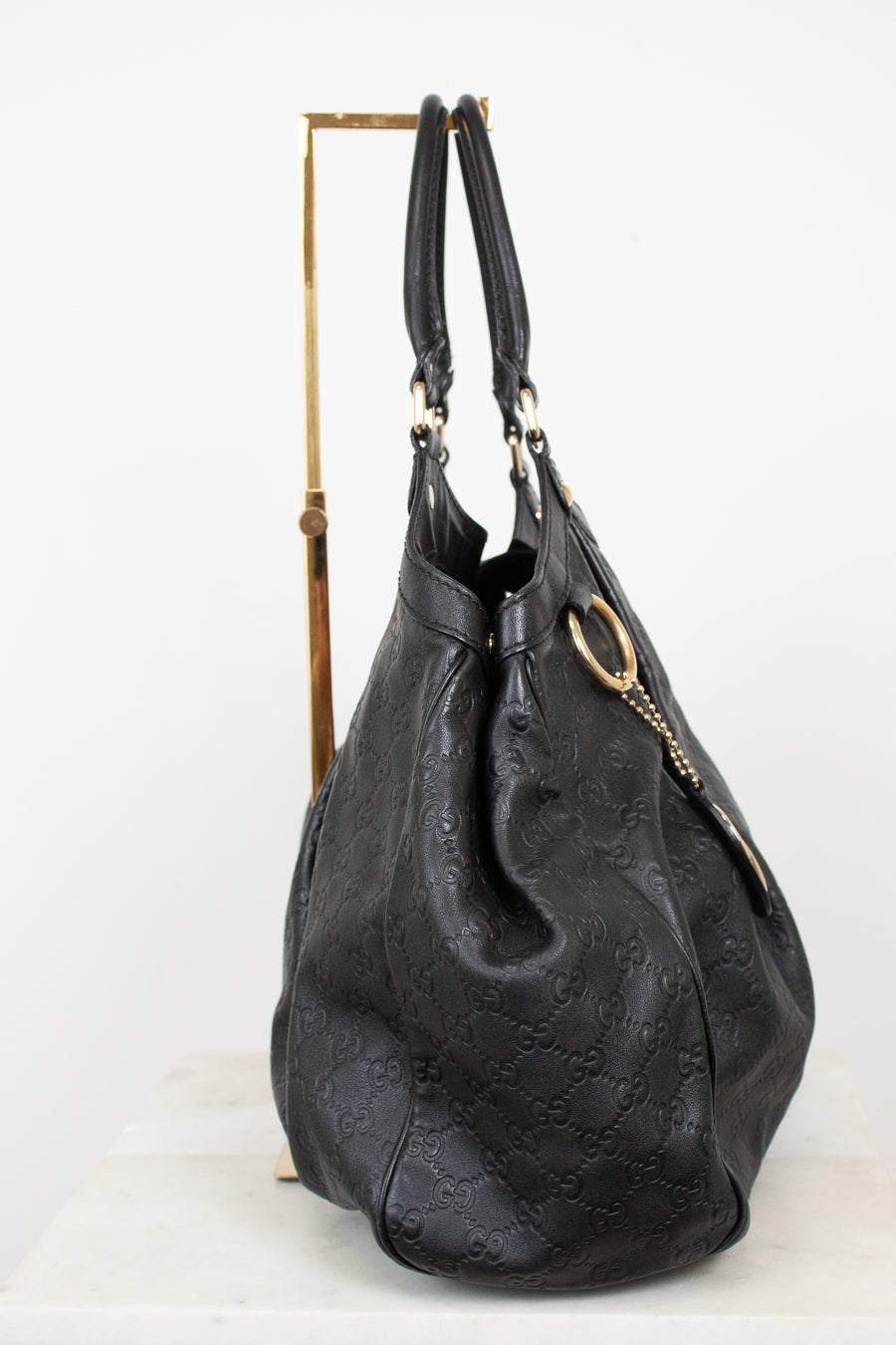 A black leather  handbag with an embossed monogram pattern, displayed on a gold metal stand against a white background.