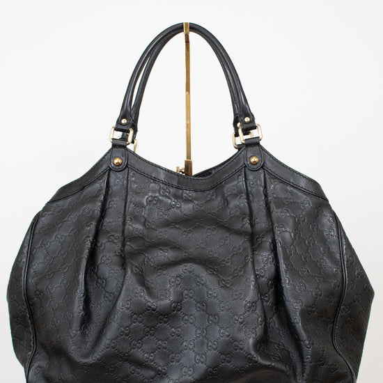 A black leather handbag with an embossed monogram pattern, featuring dual top handles and gold-tone hardware, displayed on a white pedestal against a white background.