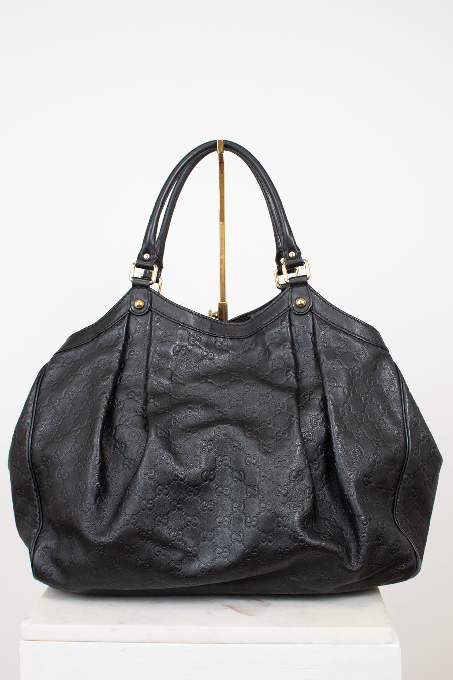 A black leather handbag with an embossed monogram pattern, featuring dual top handles and gold-tone hardware, displayed on a white pedestal against a white background.