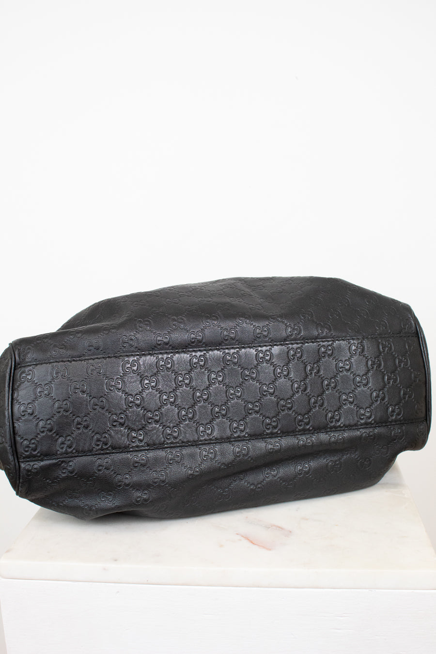 A black leather handbag with an embossed Gucci GG monogram pattern, resting on a white marble surface against a white background.