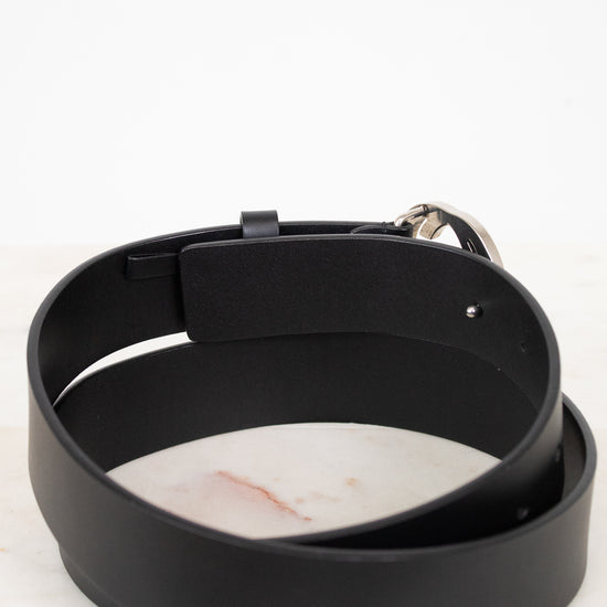 A black leather belt with a silver metal buckle, coiled on a white marble surface against a white background.