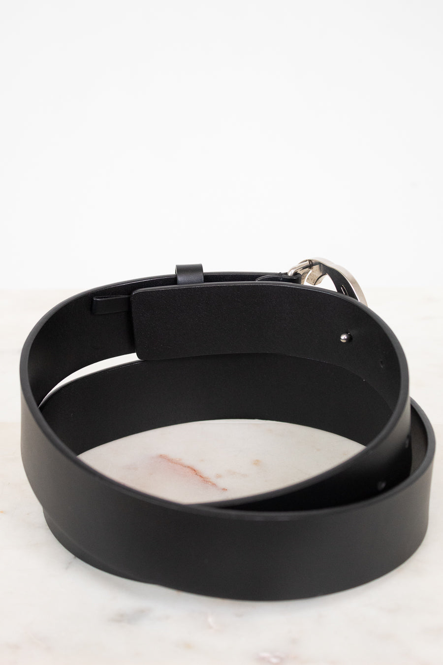 A black leather belt with a silver metal buckle, coiled on a white marble surface against a white background.