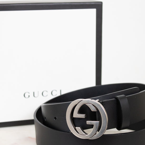A black leather Gucci belt with a silver-tone interlocking G buckle, coiled in front of its white and black Gucci box on a light marble surface.