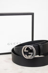 A black leather Gucci belt with a silver-tone interlocking G buckle, coiled in front of its white and black Gucci box on a light marble surface.