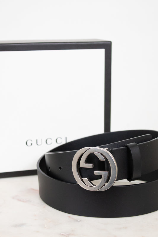 A black leather Gucci belt with a silver-tone interlocking G buckle, coiled in front of its white and black Gucci box on a light marble surface.