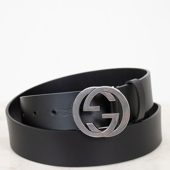 A black leather belt with a large silver-tone interlocking G buckle, coiled on a light marble surface against a white background.