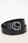 A black leather belt with a large silver-tone interlocking G buckle, coiled on a light marble surface against a white background.