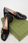 Black leather loafers with gold hardware details displayed on top of green 'GUCCI' box against a plain background