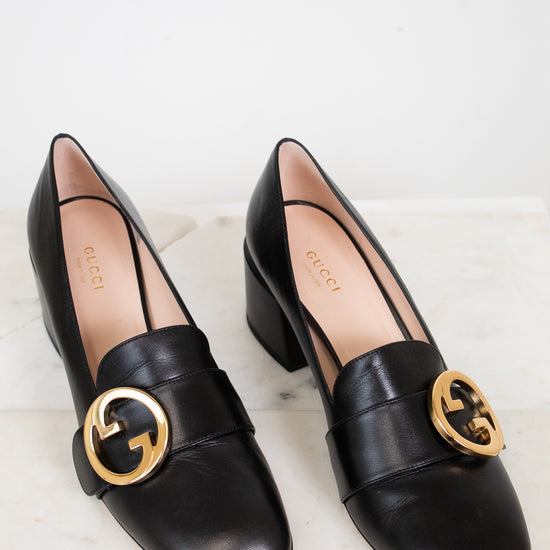 Black leather heeled loafers with gold 'GG' against a marble plain background