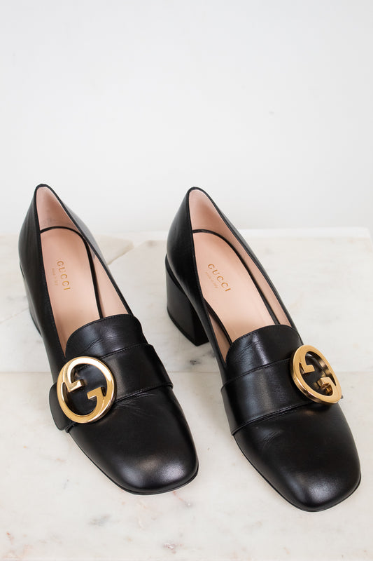 Black leather heeled loafers with gold 'GG' against a marble plain background