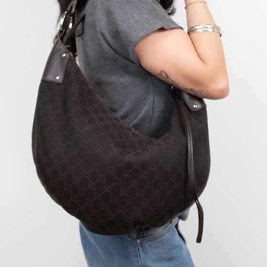 A woman wearing a gray t-shirt and blue jeans carries a dark brown Gucci hobo bag with the brand's signature GG monogram pattern on a plain light gray background.