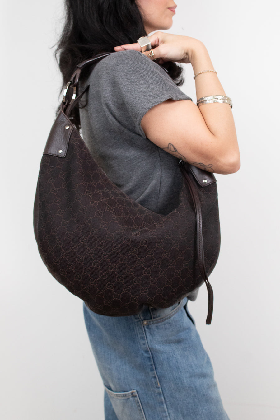 A woman wearing a gray t-shirt and blue jeans carries a dark brown Gucci hobo bag with the brand's signature GG monogram pattern on a plain light gray background.