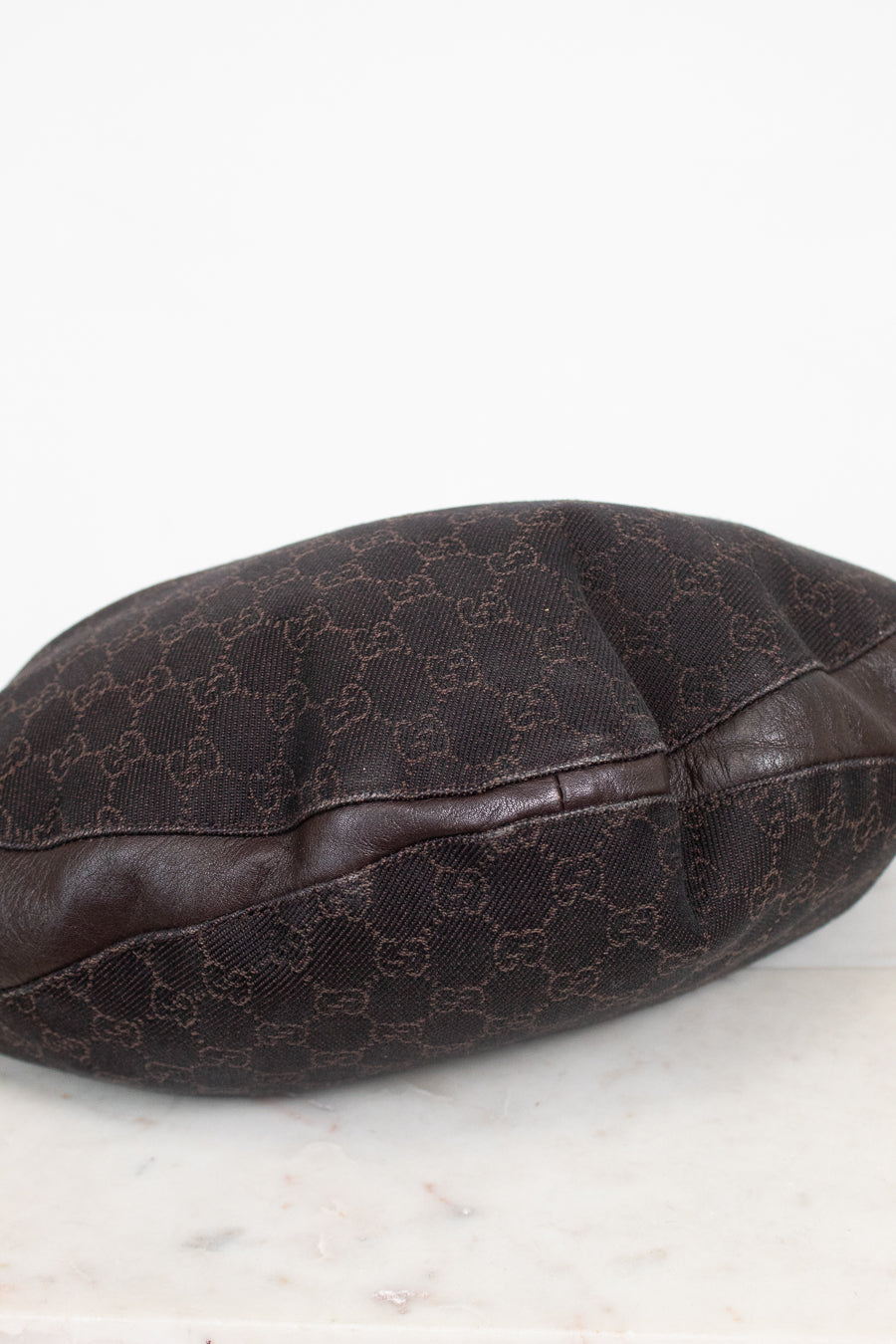 A dark brown Gucci monogram canvas and leather bag or pouch, photographed on a light marble surface against a white background.