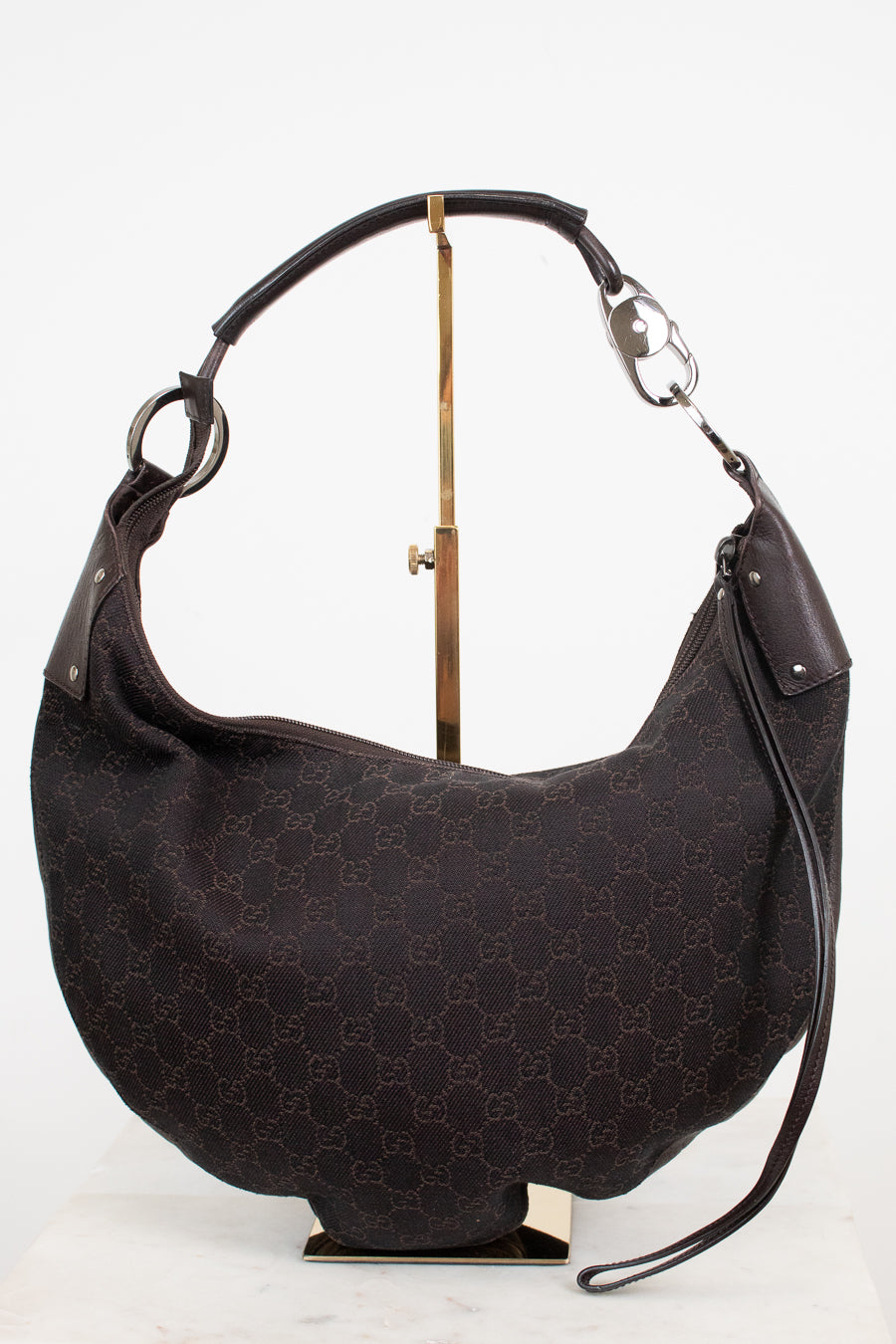 A dark brown Gucci hobo shoulder bag with the classic GG monogram canvas, displayed on a gold stand against a white background.