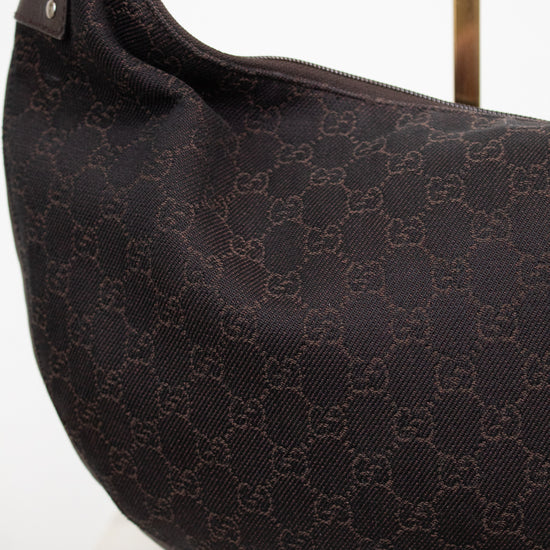 A close-up of a dark brown Gucci shoulder bag made of the brand's signature GG monogram canvas, displayed on a white surface with a gold stand visible in the background.
