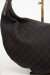 A close-up of a dark brown Gucci shoulder bag made of the brand's signature GG monogram canvas, displayed on a white surface with a gold stand visible in the background.