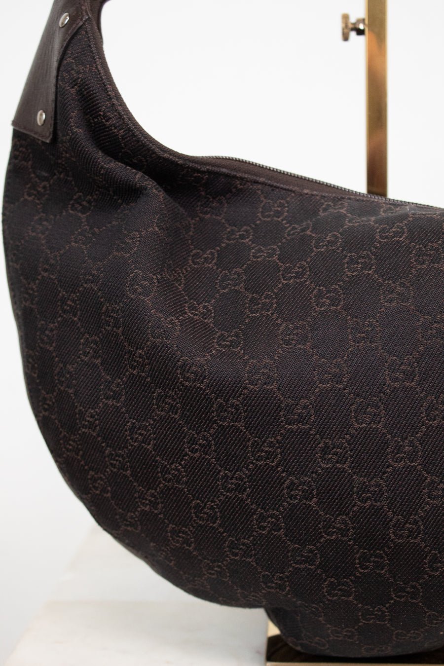 A close-up of a dark brown Gucci shoulder bag made of the brand's signature GG monogram canvas, displayed on a white surface with a gold stand visible in the background.