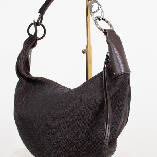 A dark brown Gucci hobo shoulder bag with the brand's signature GG monogram canvas, displayed on a gold stand against a white background.