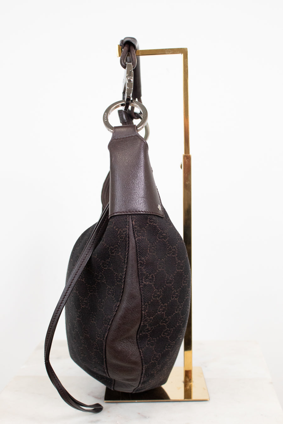 A dark brown Gucci shoulder bag with the brand's signature GG monogram canvas and leather trim, displayed on a gold metal stand against a white background.