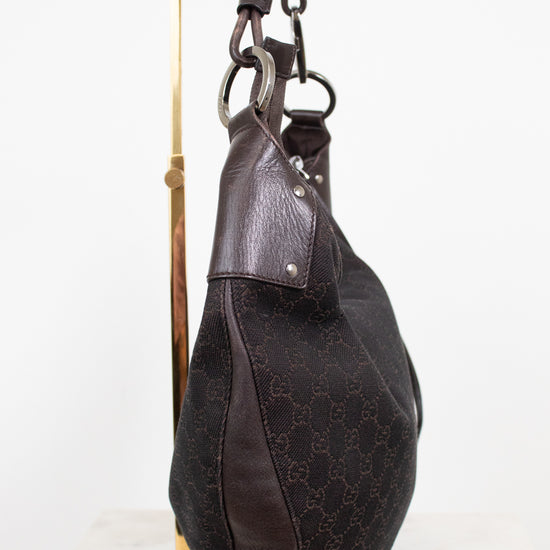 A dark brown Gucci hobo bag made of GG Supreme canvas and leather, displayed on a gold-colored metal stand against a white background.