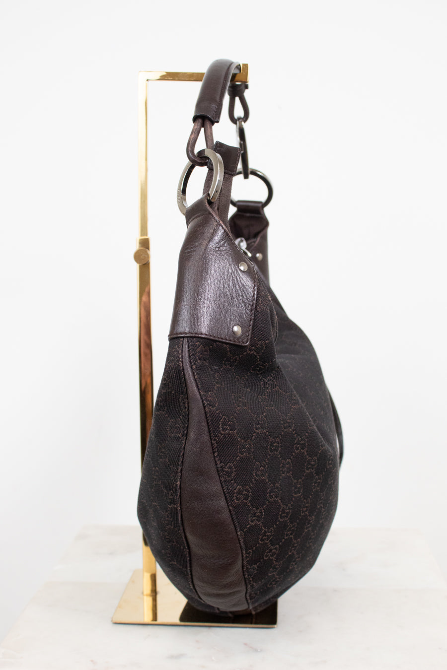 A dark brown Gucci hobo bag made of GG Supreme canvas and leather, displayed on a gold-colored metal stand against a white background.