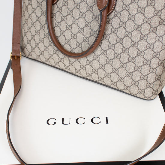 A Gucci handbag in beige and brown GG Supreme canvas with brown leather handles and shoulder strap, displayed on top of a white Gucci box.