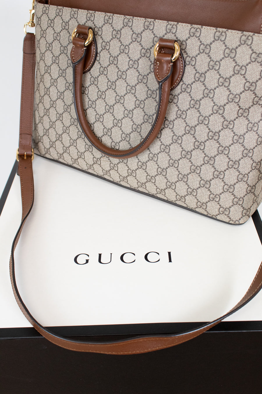 A Gucci handbag in beige and brown GG Supreme canvas with brown leather handles and shoulder strap, displayed on top of a white Gucci box.