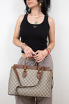 A woman wearing a black tank top and gray pants holds a beige and brown Gucci GG Supreme tote bag with gold-tone hardware against a plain white background.