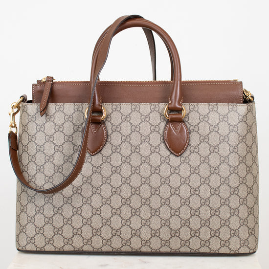 A Gucci GG Supreme tote bag in beige and brown monogram canvas with brown leather handles and trim, displayed on a white marble block against a white background.
