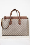 A Gucci GG Supreme tote bag in beige and brown monogram canvas with brown leather handles and trim, displayed on a white marble block against a white background.