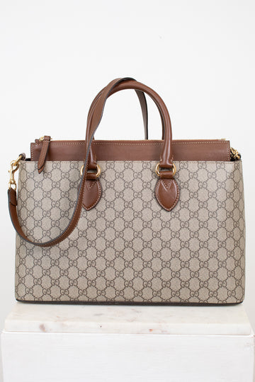 A Gucci GG Supreme tote bag in beige and brown monogram canvas with brown leather handles and trim, displayed on a white marble block against a white background.