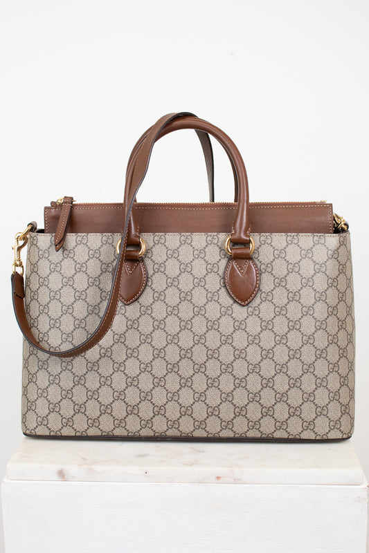 A Gucci GG Supreme tote bag in beige and brown monogram canvas with brown leather handles and trim, displayed on a white marble block against a white background.