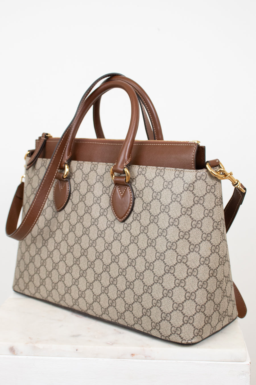 A Gucci GG Supreme handbag in beige and brown canvas with brown leather handles and trim, shown on a white marble surface against a white background.