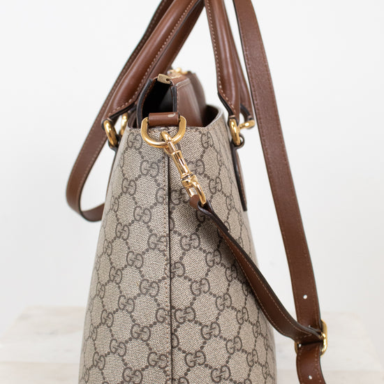 A Gucci handbag in beige GG Supreme canvas with brown leather trim and gold-tone hardware, shown on a light neutral background.