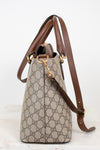 A Gucci handbag in beige GG Supreme canvas with brown leather trim and gold-tone hardware, shown on a light neutral background.