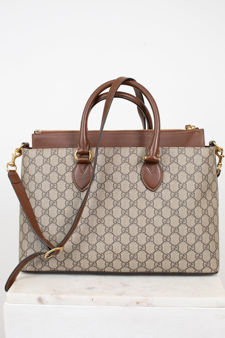 A Gucci monogram canvas handbag with brown leather handles and trim, displayed on a white marble block against a white background.
