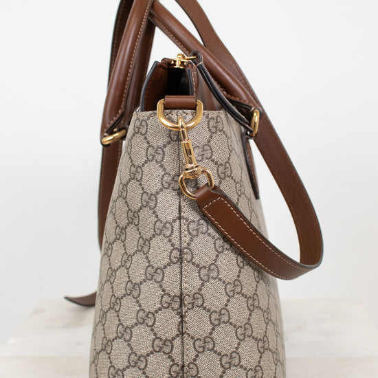 A Gucci handbag in beige and brown GG Supreme canvas with brown leather trim and gold-tone hardware, shown from the side against a plain white background.