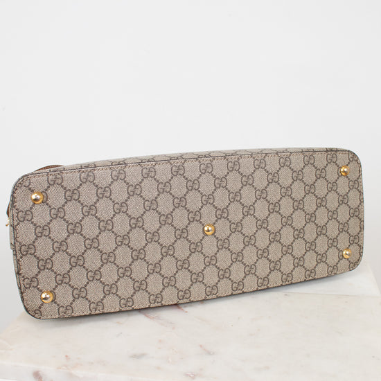 A Gucci shoulder bag in beige and brown GG Supreme canvas with gold-tone studs, resting on a white marble surface against a white background.