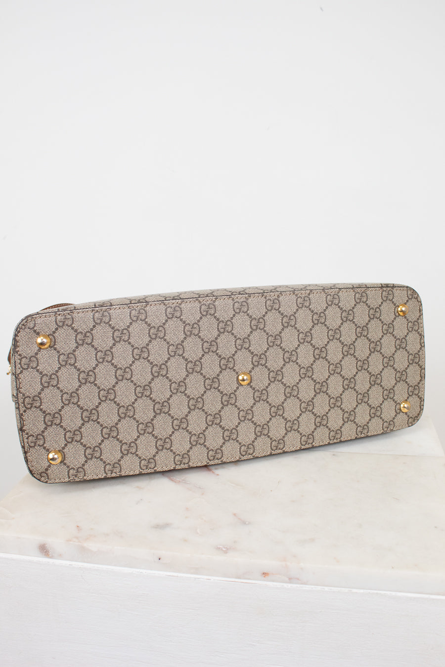 A Gucci shoulder bag in beige and brown GG Supreme canvas with gold-tone studs, resting on a white marble surface against a white background.